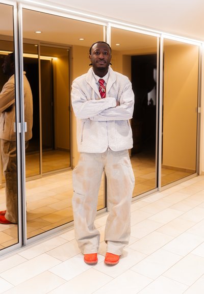 Hombre de pie con los brazos cruzados, vistiendo un chaleco de forro polar blanco, camisa a rayas, corbata roja, pantalones beige y zapatos de color naranja brillante en un pasillo moderno.