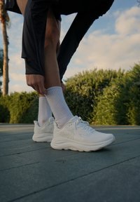 Bele športne čevlje z teksturiranim zgornjim delom in debelim podplatom, v kombinaciji z rebrastimi belimi nogavicami, na lesenem površju, obdanim z zelenjem.