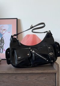 Czarna torebka z lakierowanej skóry o zaokrąglonym kształcie. Posiada wiele kieszeni na zamek i na klapę, srebrne okucia oraz regulowany pasek.