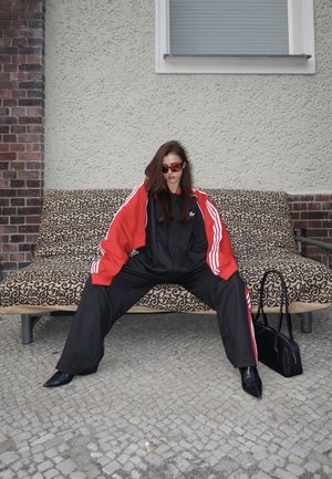 Mujer con chándal rojo y negro y gafas de sol sentada con las piernas abiertas en un banco con estampado al aire libre, bolso negro en el suelo cerca.