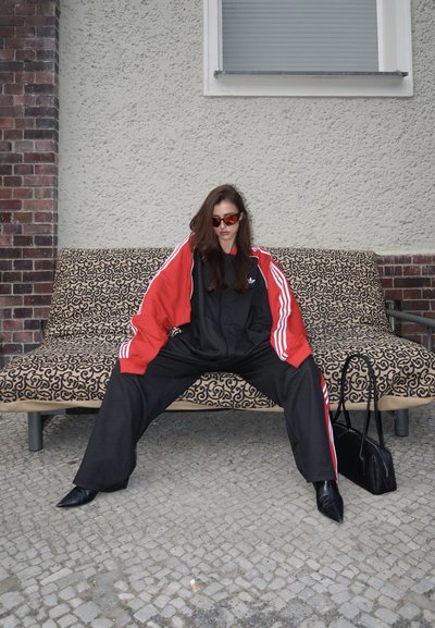 Mujer con chándal rojo y negro y gafas de sol sentada con las piernas abiertas en un banco con estampado al aire libre, bolso negro en el suelo cerca.