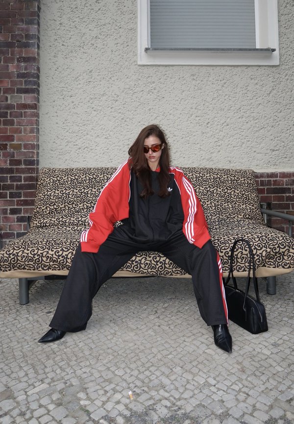 Kobieta w czerwonym i czarnym dresie oraz okularach przeciwsłonecznych siedząca na szeroko rozstawionych nogach na wzorzystej ławce na zewnątrz, czarna torebka leżąca obok na ziemi.