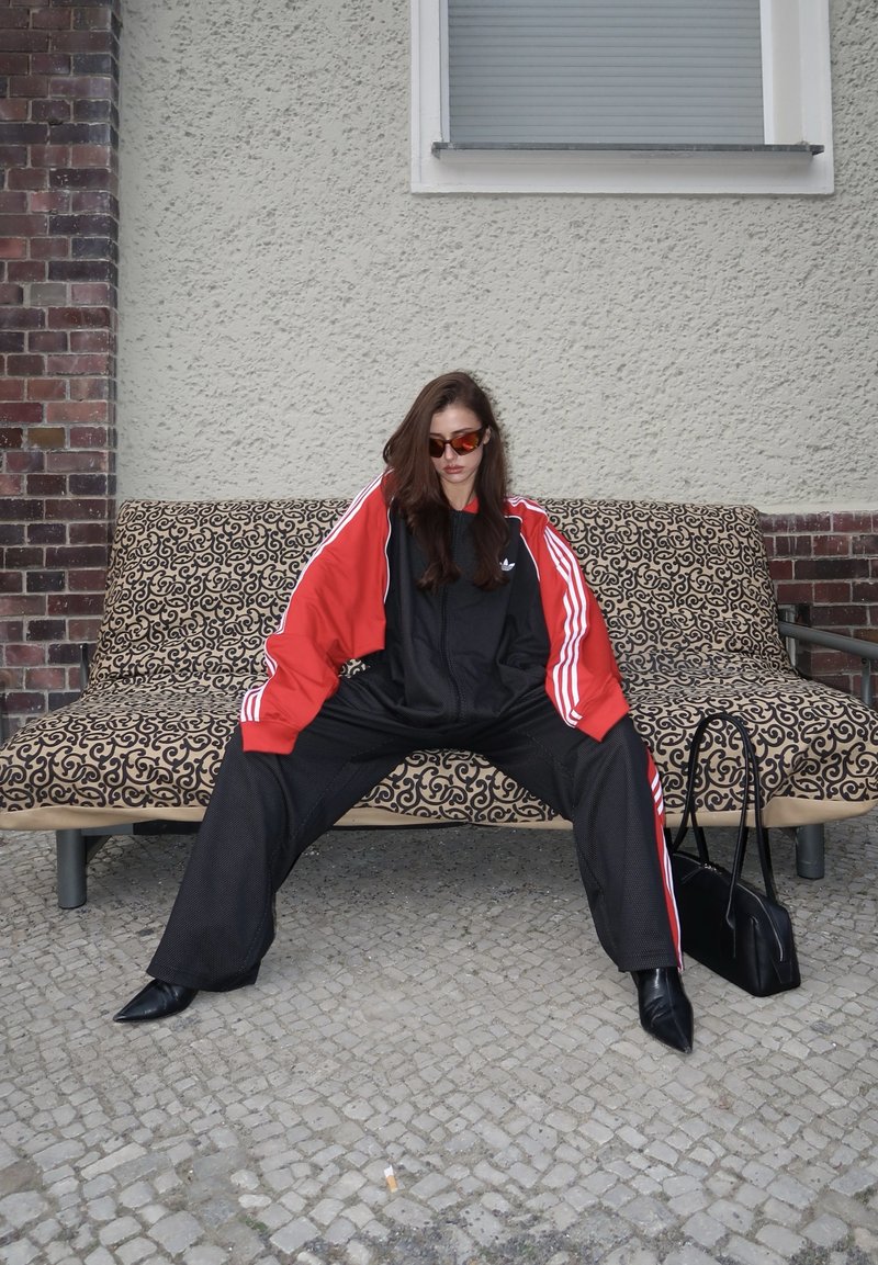 Γυναίκα με κόκκινη και μαύρη αθλητική φόρμα και γυαλιά ηλίου κάθεται με ανοιχτά πόδια σε παγκάκι με σχέδια σε εξωτερικό χώρο, μαύρη τσάντα στο έδαφος κοντά της.