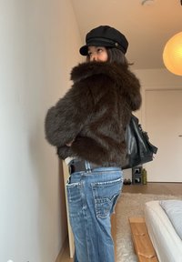 Femme portant une veste en fourrure foncée, une casquette noire, un jean bleu et un sac à dos noir, debout à l'intérieur près d'un lit et d'une lampe.