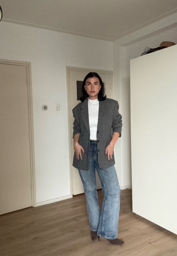 Une femme présente un blazer à carreaux, un haut blanc et un jean à jambes larges dans une pièce.