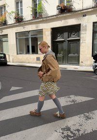 Brązowa oversize'owa kurtka na szarej bluzie z kapturem, kratkowana mini spódnica w zielono-żółte wzory, szare skarpetki do kolan, brązowe buty z kwiatowymi detalami.