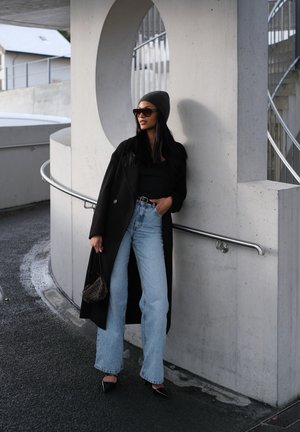Abrigo largo negro sobre una blusa negra, jeans de pierna ancha azul claro, tacones negros afilados, gorro gris y gafas de sol. Se sostiene un bolso estampado.