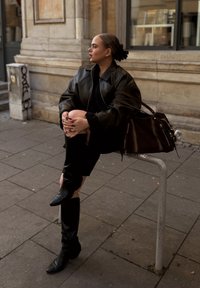 Žena v černé kožené bundě a botách sedí na kovovém schůdku na chodníku ve městě, má ruce položené na kolenou, hledí na stranu.