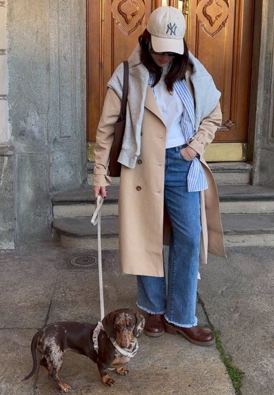 Abrigo trench beige sobre un sudadera gris, camisa blanca y vaqueros azules. Botas marrones hasta el tobillo. Correa sujeta a un pequeño dachshund marrón.