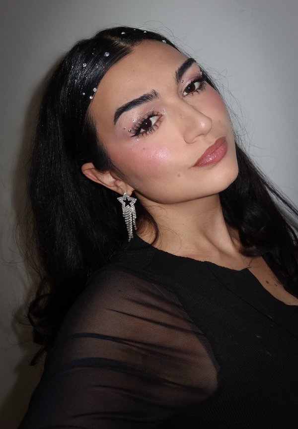 Chemise longue à manches longues noire transparente, boucles d'oreilles étoiles argentées avec perles en cascade, maquillage pailleté subtil et cheveux foncés texturés ornés de gemmes décoratives.
