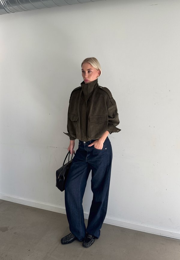 Mujer rubia con una chaqueta oscura de gran tamaño, vaqueros anchos oscuros y zapatos negros, llevando un bolso negro, de pie contra una pared lisa.