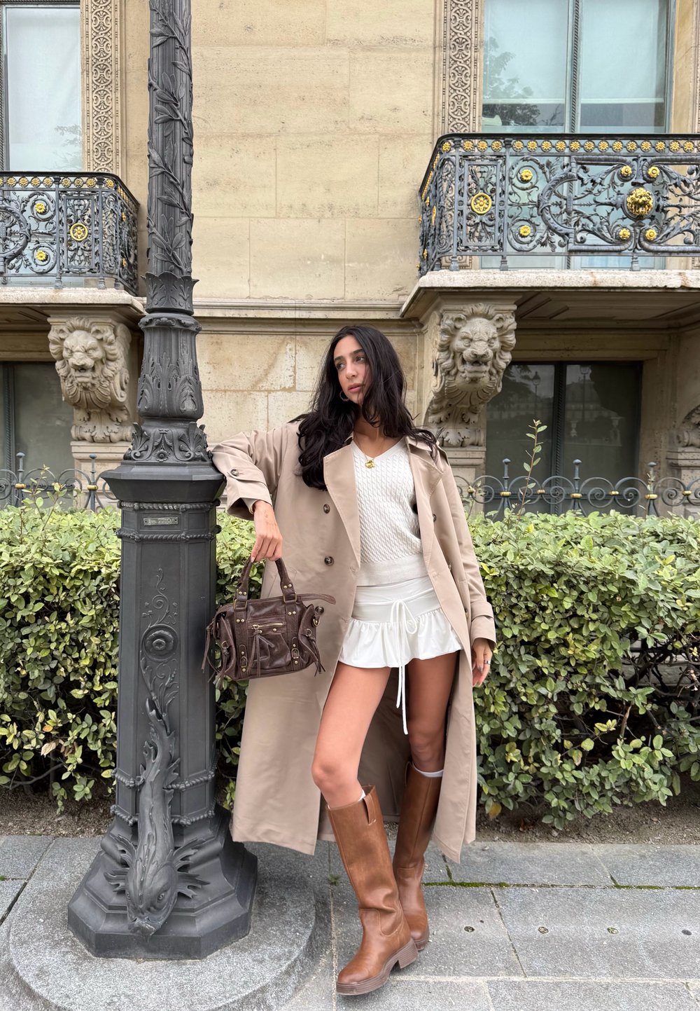 Donna in un trench beige, maglione bianco e gonna, stivali marroni e borsa, appoggiata a un lampione decorativo davanti a una facciata di edificio ornata.