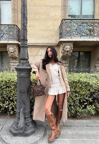 Donna in un trench beige, maglione bianco e gonna, stivali marroni e borsa, appoggiata a un lampione decorativo davanti a una facciata di edificio ornata.