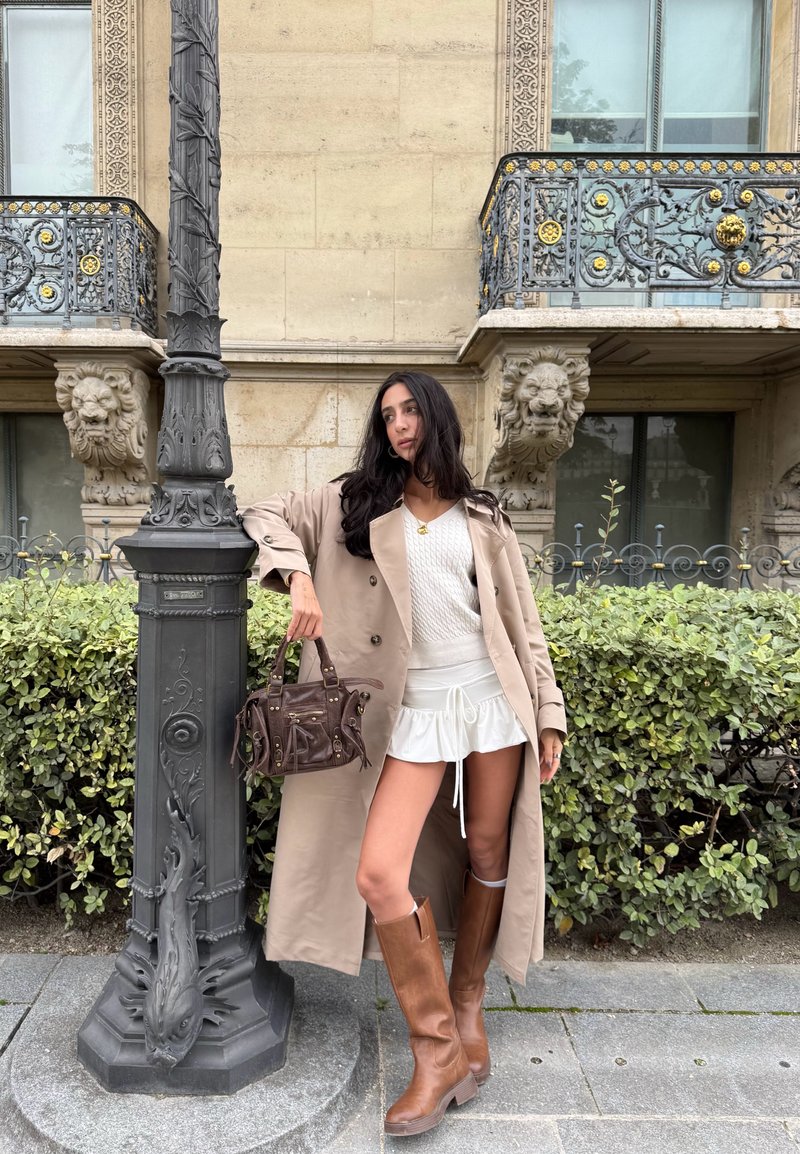 Mulher com um trench coat bege, suéter branco e saia, botas castanhas e uma bolsa, apoiada num candeeiro decorativo em frente a uma fachada de edifício ornamentada.
