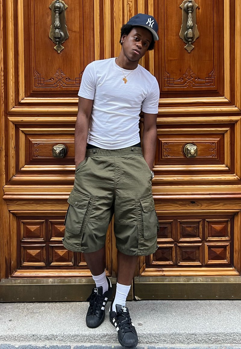 T-shirt blanc, shorts cargo verts olive avec poches latérales, baskets noires avec accents blancs, et une casquette de baseball bleue. Fond en porte en bois.