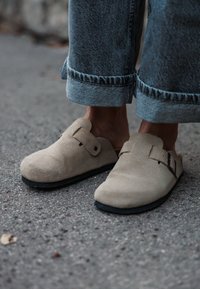 Sabots en suédine beige avec semelles noires plates, dotés d'un bout arrondi et d'une sangle réglable. Portés avec un jean en denim bleu clair retroussé.
