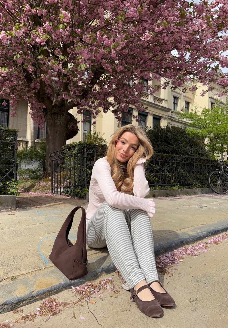 Νεαρή γυναίκα με μακριά ξανθά μαλλιά κάθεται στο πεζοδρόμιο κάτω από ροζ ανθισμένο δέντρο, φοράει ριγέ παντελόνια, ανοιχτό πουλόβερ και σκούρα παπούτσια, δίπλα σε καφέ τσάντα.