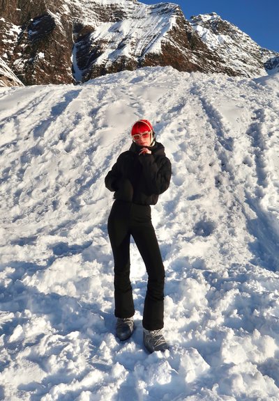 Chaqueta de nieve aislante negra y pantalones, botas de nieve plateadas, cabello rojo, gafas de sol blancas de gran tamaño, de pie sobre la nieve con montañas rocosas de fondo.