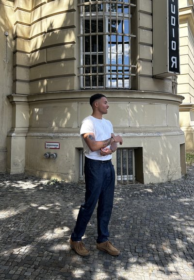 Camisa blanca de mangas cortas, pantalones de mezclilla azul oscuro y zapatos marrones. De pie sobre adoquines con un edificio que presenta ventanas con rejas al fondo.