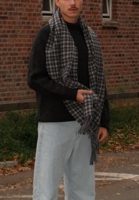 Schwarzer Strickpullover, hellblaue Jeans und ein kariertes graues Schal mit Fransen, lässig mit einer Hand gehalten. Ziegelstein-Hintergrund.
