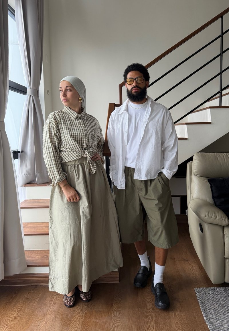 Femme portant un foulard et une longue jupe et homme avec barbe en short et chemise blanche debout à l'intérieur près d'un escalier et d'une fenêtre.