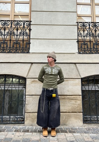 Top de manga larga verde oliva con estampado decorativo blanco, combinado con jeans anchos negros, zuecos color tostado y un accesorio amarillo colgando del cinturón.