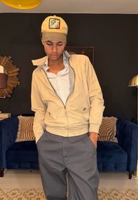 Jeune homme portant une casquette beige, une veste claire et un pantalon gris, debout avec les mains dans les poches dans un salon aux murs sombres et avec un canapé bleu.