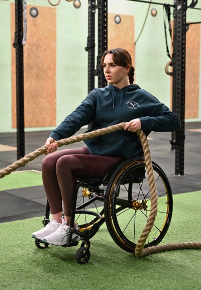 Mujer en silla de ruedas haciendo ejercicio con cuerda de batalla en el gimnasio, vestida con ropa deportiva y concentrada en su entrenamiento.