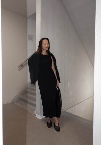 Vestido maxi de punto acanalado negro sin mangas, combinado con una chaqueta negra caída sobre los hombros y un bolso negro estructurado.