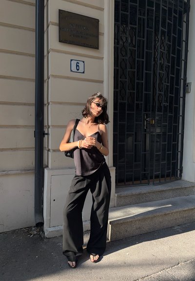 Camiseta de tirantes finos negra, pantalones negros holgados y sandalias planas. De pie contra un edificio con una puerta decorativa y señalización.