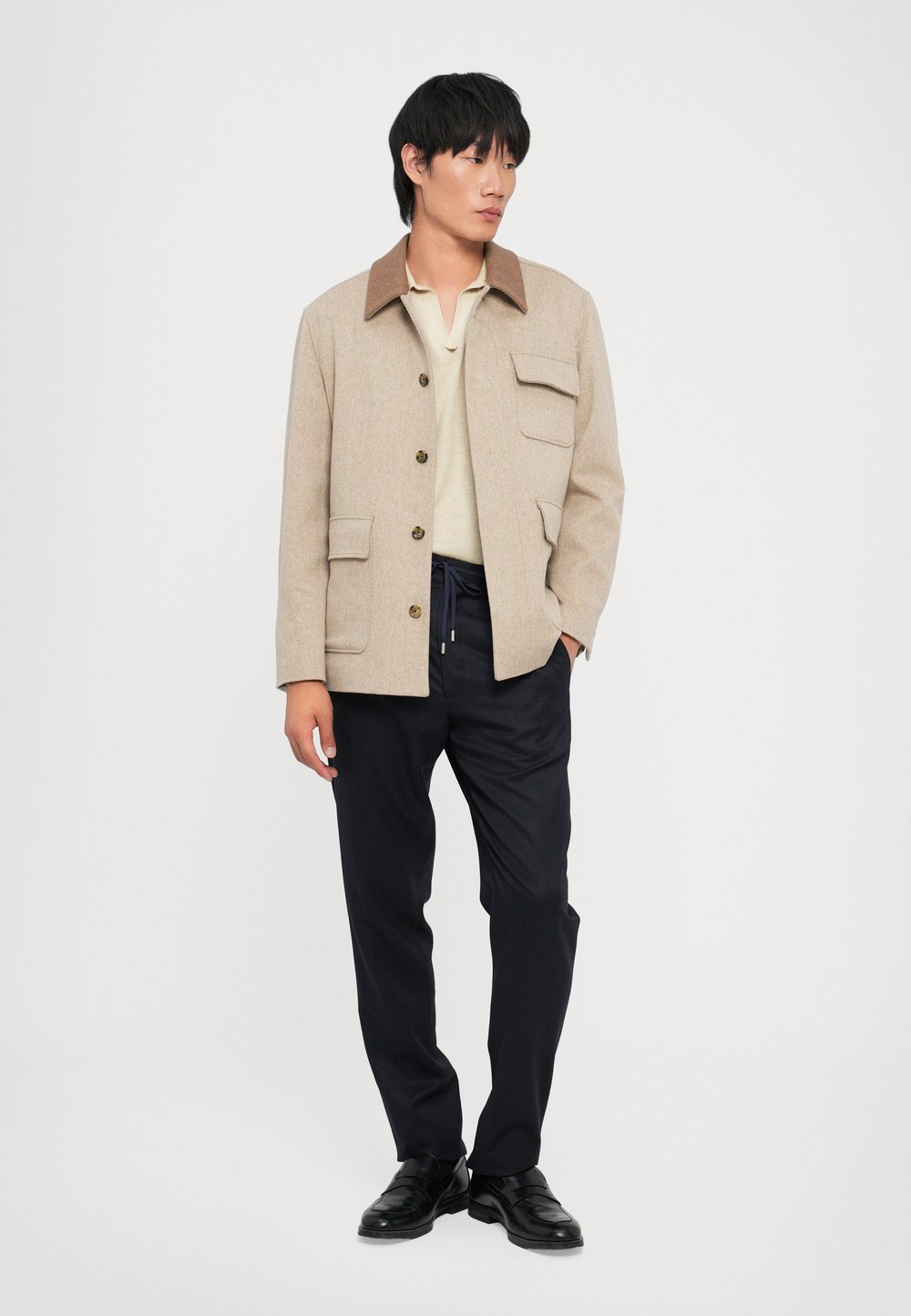 Beige ullblandingsjakke med brun krage, frontlukking med knapper og to klaffelommer. Brukt over en lys polo og marineblå bukser.