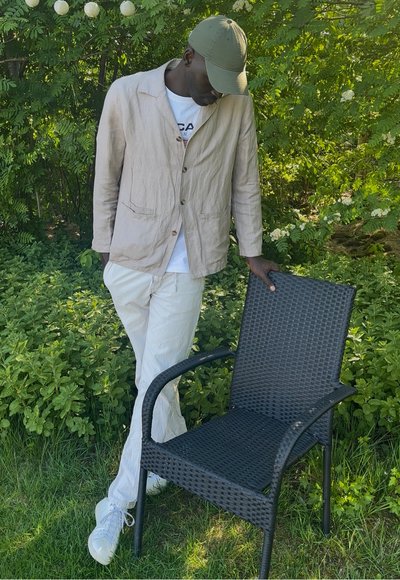 Chaqueta de lino beige claro sobre una camiseta blanca, combinada con pantalones claros. Una silla de exterior tejida negra se encuentra cerca, contra un fondo de follaje verde.