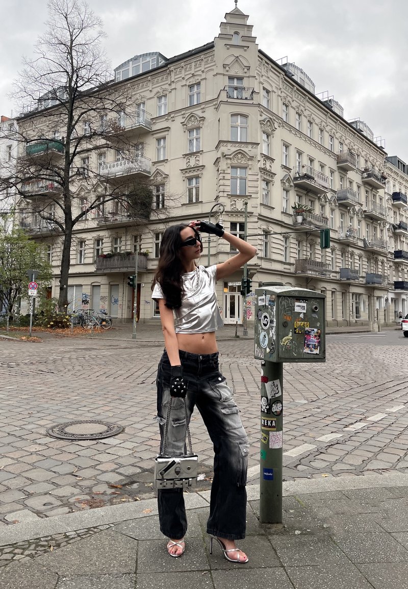 Μεταλλική κομμένη μπλούζα, φθαρμένο γκρι-μαύρο cargo παντελόνι, ασημένιες ψηλοτάκουνες σανδάλια, γάντια χωρίς δάχτυλα, κρατώντας μια μεταλλική τσάντα, σε αστικό δρόμο.