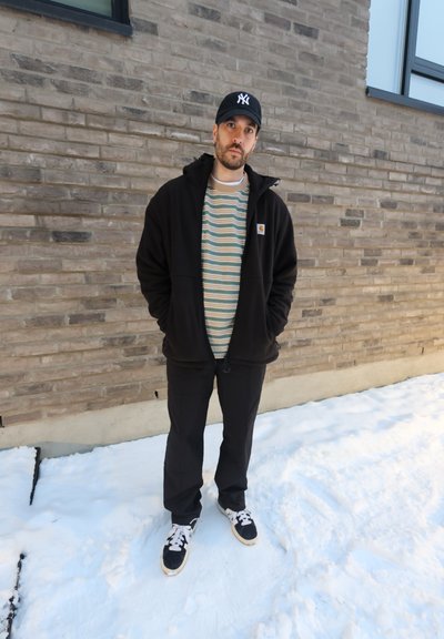 Hombre con chaqueta negra y gorra está de pie sobre la nieve frente a una pared de ladrillo con ventanas, con las manos en los bolsillos, mirando a la cámara.