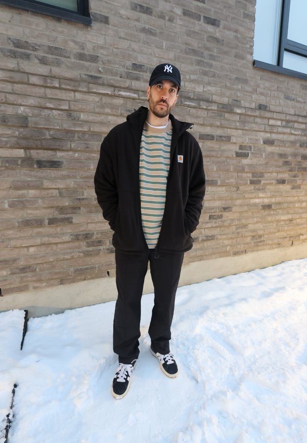 Muž v čiernej bunde a čiapke stojí na snehu pred tehlovou stenou s oknami, ruky v vreckách, pozerá sa do kamery.