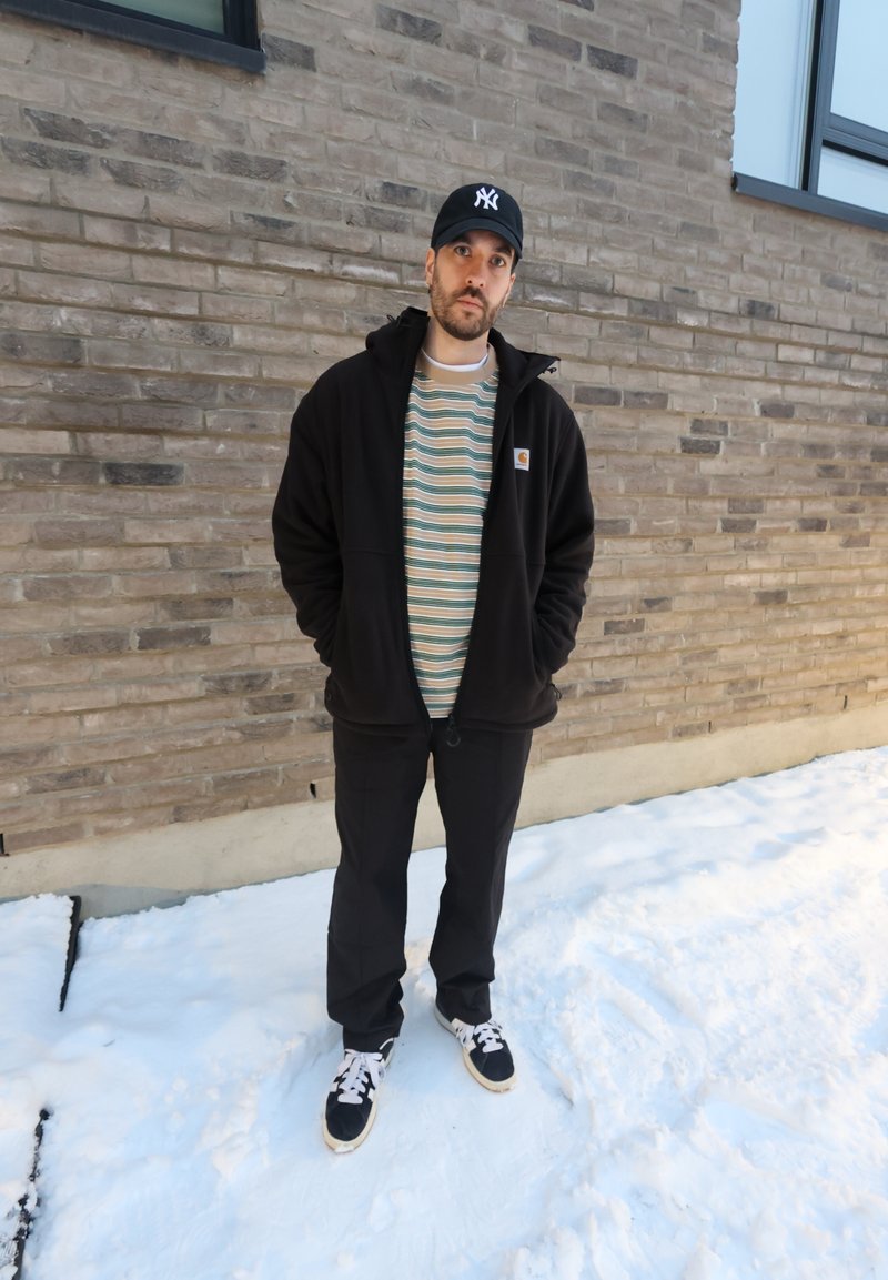 Homme portant une veste noire et une casquette, debout sur la neige devant un mur de briques avec des fenêtres, les mains dans les poches, regardant la caméra.