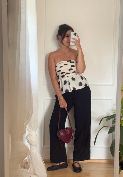 Top blanco sin tirantes con manchas negras y cintura peplum. Pantalones negros de tiro alto con una textura suave. Bolsa en forma de corazón en color burdeos y bailarinas negras.