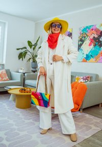 Femme en manteau blanc, chapeau jaune, écharpe rouge, tenant un sac rayé arc-en-ciel, debout dans un salon coloré avec de l'art moderne et des plantes.
