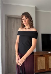 Sieviete, kas valkā melnu topu ar atvērtām pleciem, bordo krāsas svārkus un zelta rotaslietas, stāv telpās pie koka skapīša un televizora.