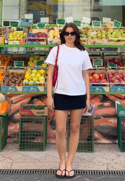 Mujer con camiseta blanca, falda negra y gafas de sol está delante de un puesto de frutas con manzanas, peras y aguacates.