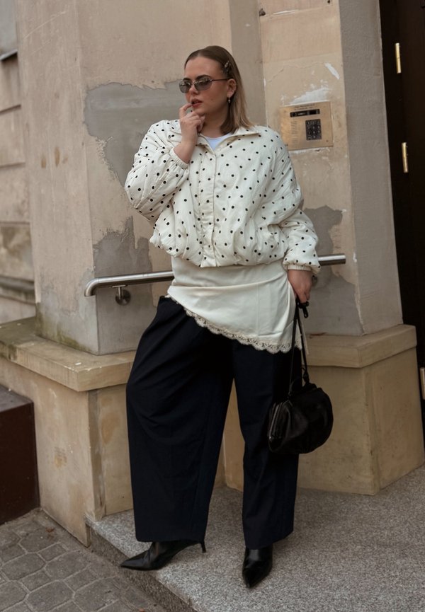 Femeie purtând geacă matlasată cu buline, pantaloni largi negri, cizme negre ascuțite, ținând o geantă neagră, stând lângă o clădire din piatră bej.