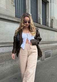 Καφέ δερμάτινο μπουφάν, λευκό μπλουζάκι με κορδόνι, και ανοιχτό μπεζ λινά παντελόνια. Τα αξεσουάρ περιλαμβάνουν μικρές στρογγυλές αντηλιάδες και κρεμαστές αλυσίδες.