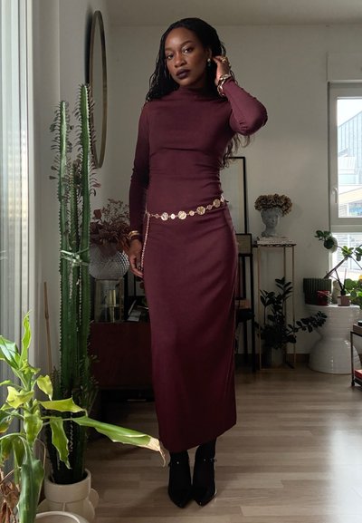 Vestido burdeos ajustado de manga larga con cuello alto simulado y cinturón metálico dorado en la cintura. Zapatos de tacón puntiagudos negros y accesorios mínimos. Entorno interior.