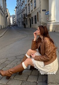 Braune Lederjacke, weißes Spitzenkleid mit gewelltem Saum, knielange braune Stiefel mit weißen Akzenten, sitzt auf einer Kopfsteinpflasterstraße.
