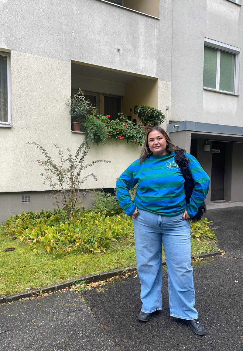 Camisa de mangas compridas às riscas em azul petróleo e azul marinho com um gráfico de número, combinada com jeans de perna larga em azul claro e sapatos pretos, de pé ao ar livre.