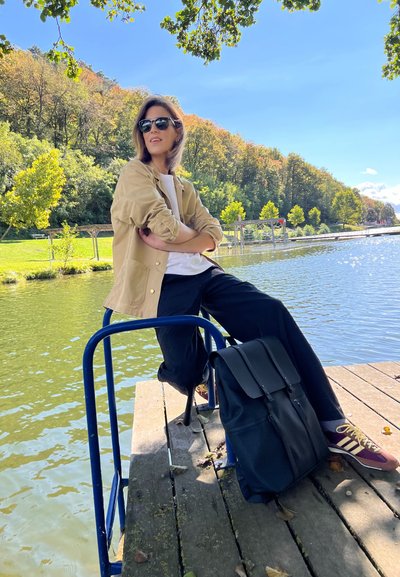 Mochila negra con tapa, chaqueta beige sobre camiseta blanca, pantalones negros, sentado en un muelle de madera junto al agua verde, gafas de sol, zapatillas casuales.