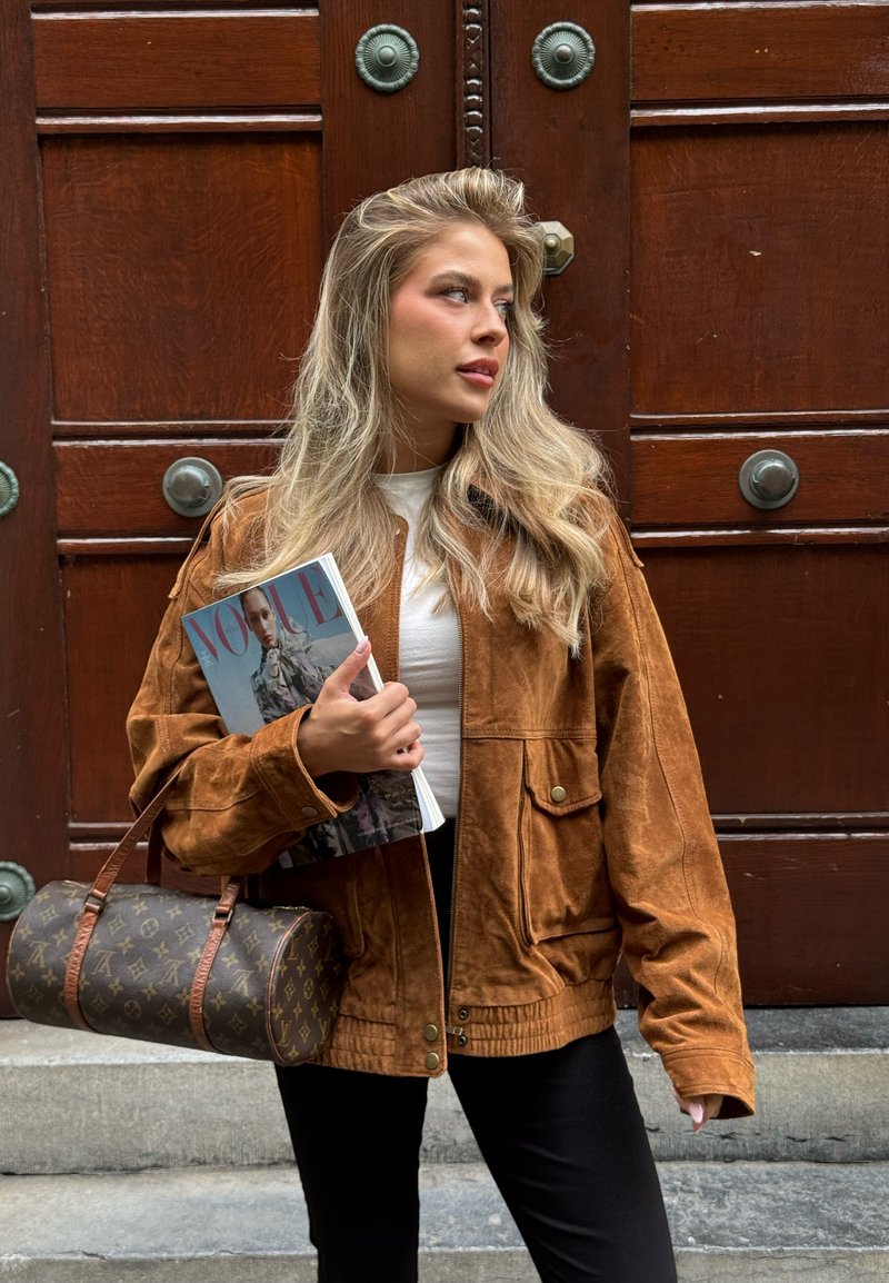 Veste en daim marron au style décontracté, dotée de grandes poches et de boutons. Tient un magazine et un sac fourre-tout marron monogrammé.
