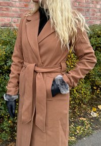 Camel wool-blend coat with a wrap design, featuring a belted waist, wide lapels, and fur-trimmed cuffs. Black gloves complete the look.
