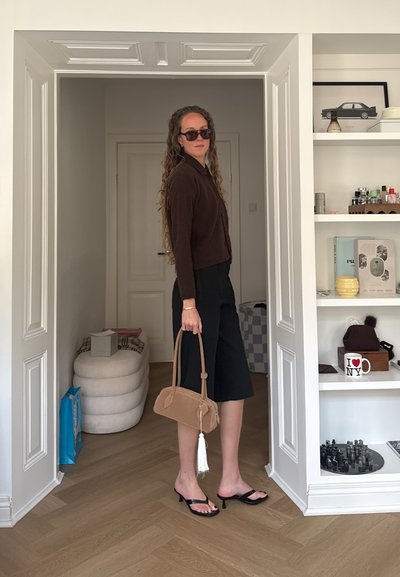 Mujer con cabello largo y rizado que lleva gafas de sol, cárdigan marrón, culottes negros y sandalias negras de tacón sosteniendo un bolso marrón claro en el interior.