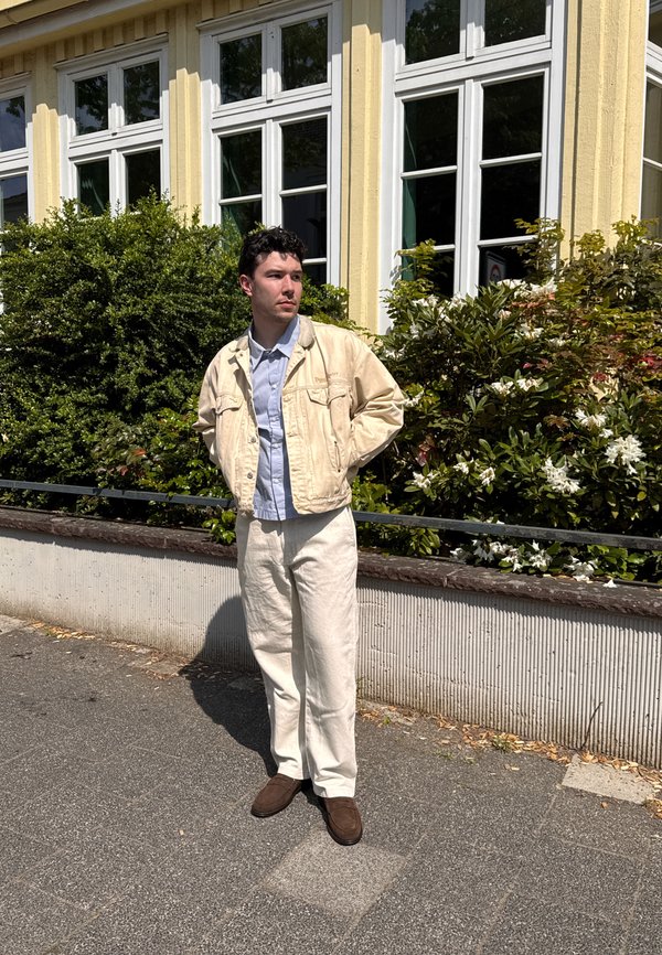 Homme portant une veste beige et un pantalon avec des chaussures marron se tient sur le trottoir à côté de buissons verts et d'un bâtiment jaune avec des fenêtres blanches.